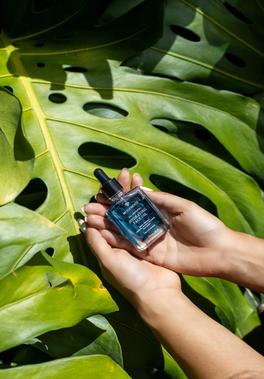 Hand holding a small bottle of blue liquid against a backdrop of large green leaves